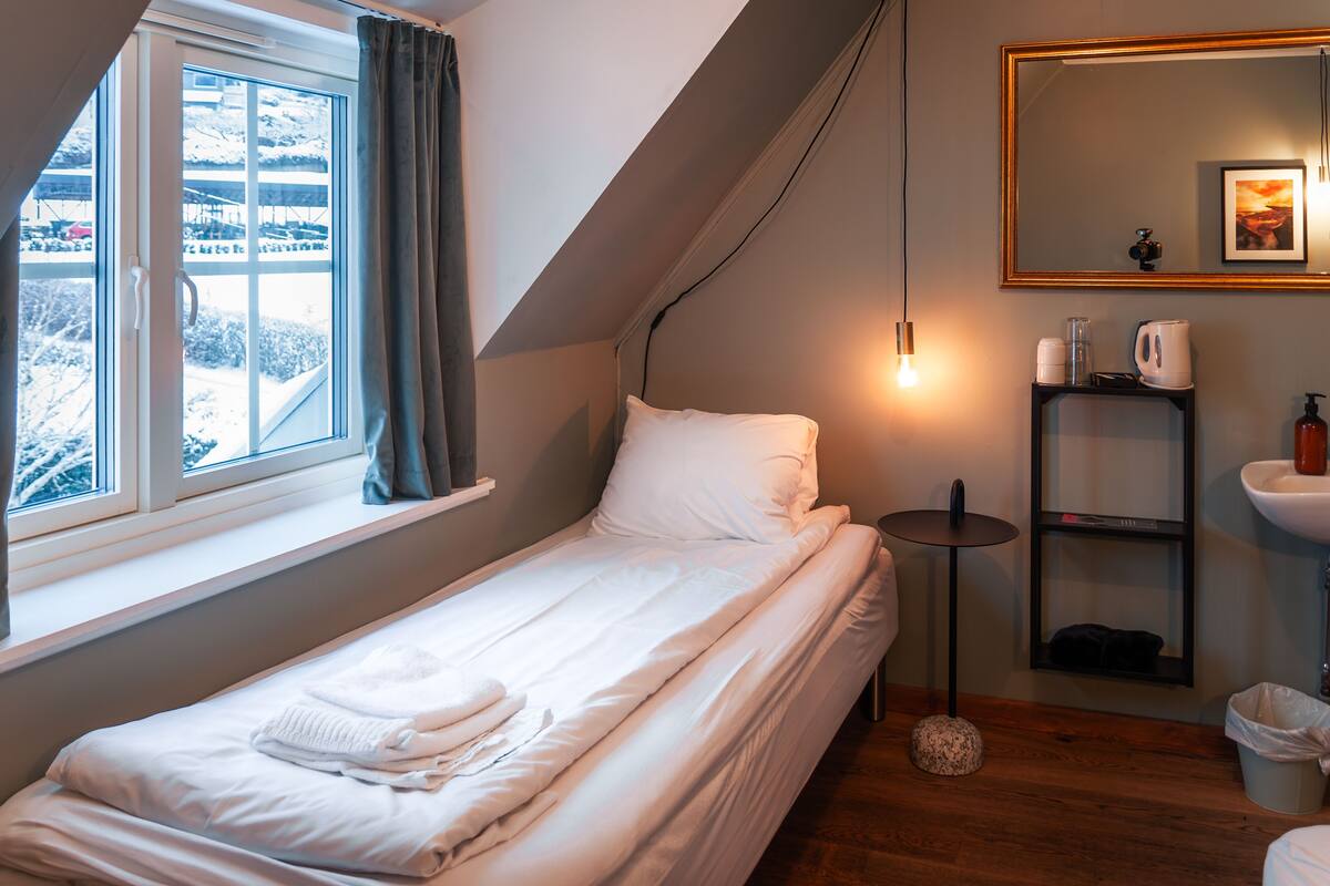 A small, neatly made bed with white linens, a bedside table with a lamp, a mirror, and a shelf with toiletries.