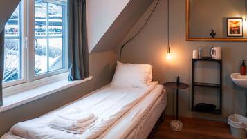 Un letto piccolo e ben rifatto con lenzuola bianche, un comodino con una lampada, uno specchio e uno scaffale con articoli da toilette.