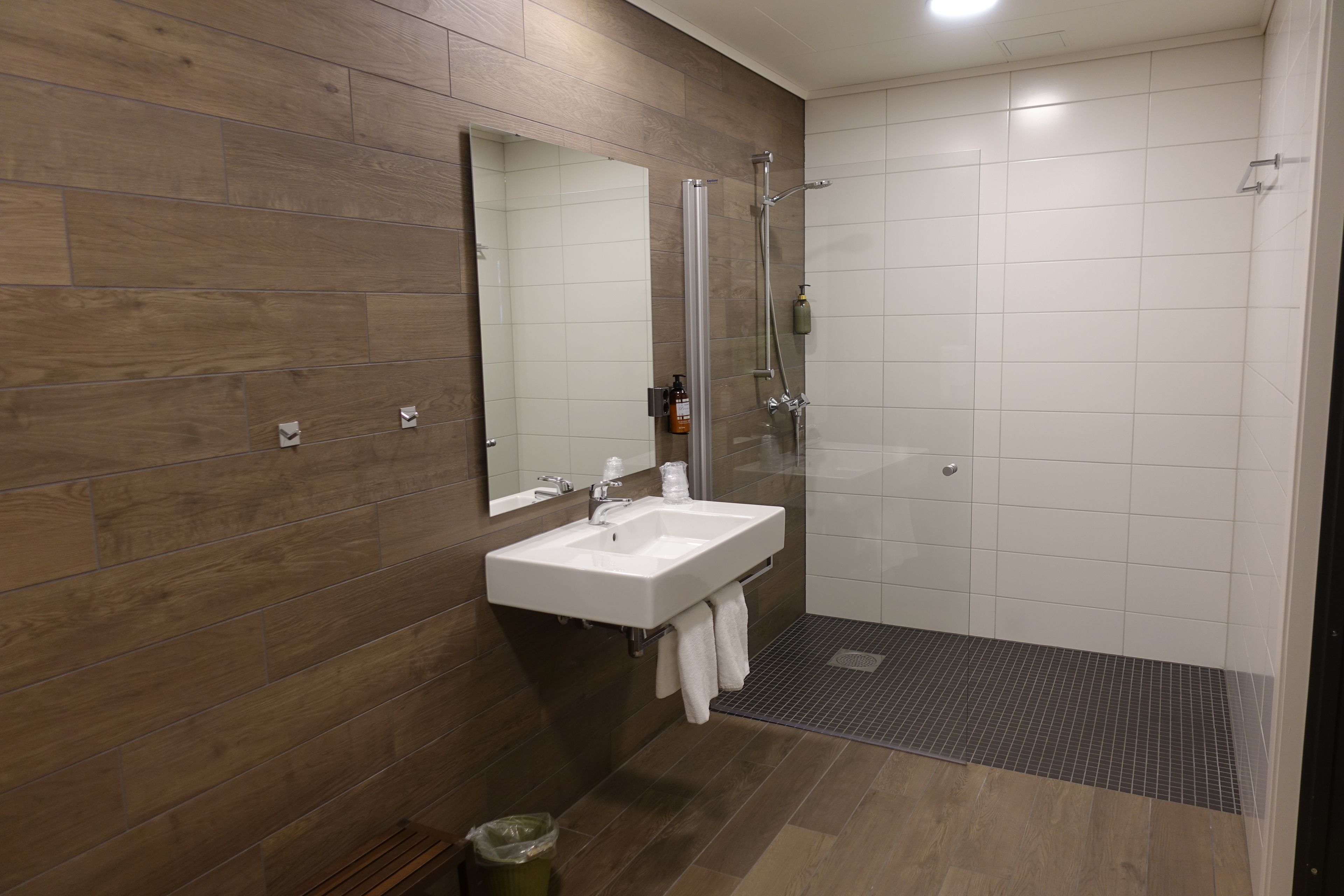 A modern bathroom with a wooden wall, a white sink, a glass shower enclosure, and a tiled floor.