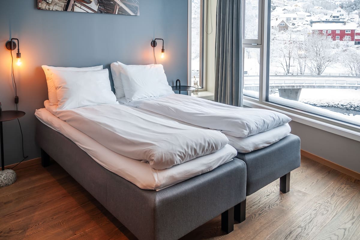 A double bed with white linens, a grey bench, and a nightstand with a lamp.