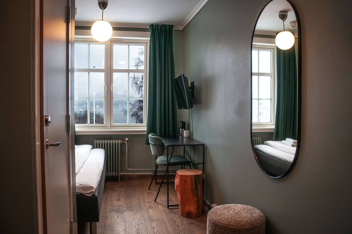 A compact hotel room with a bed, a desk with a chair, a round wooden stool, a mirror, and a view of the outdoors through a window with curtains.
