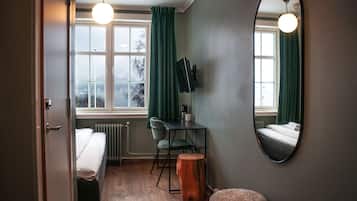 Une chambre d’hôtel de taille réduite, comprenant un lit, un bureau avec une chaise, un tabouret rond en bois, un miroir et une vue sur l’extérieur par une fenêtre munie de rideaux.