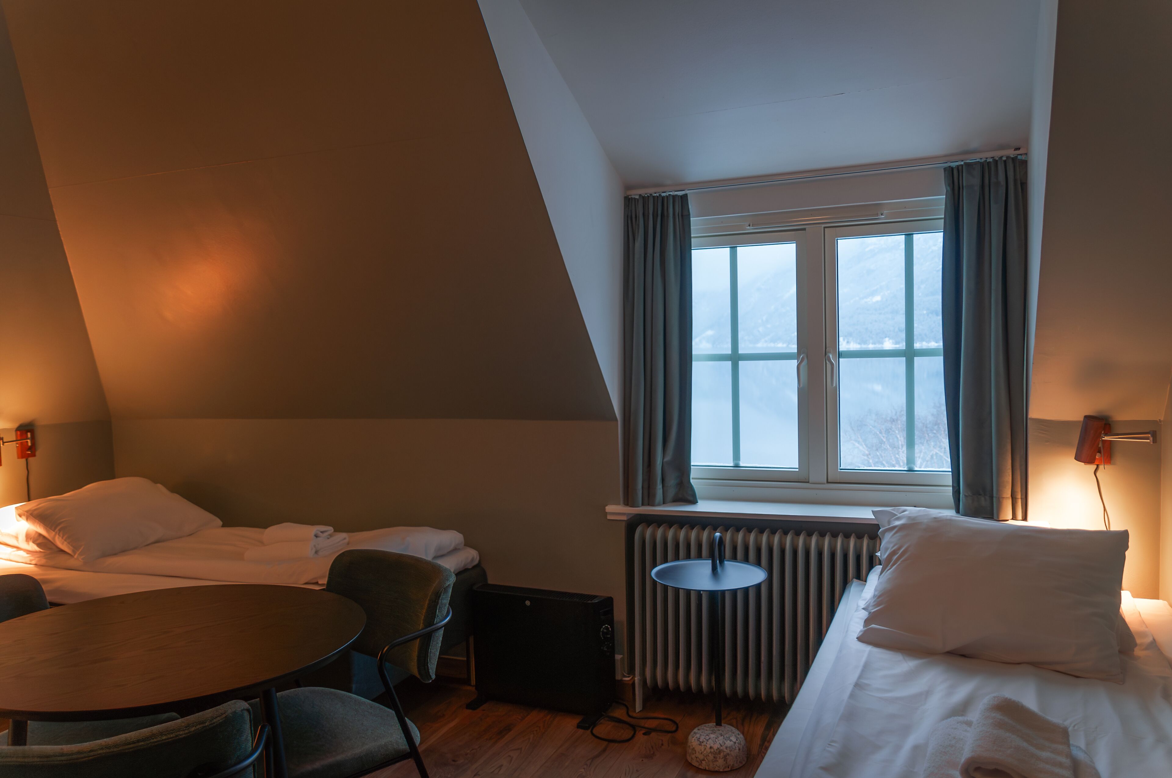 A hotel room with a bed, a round table, a chair, a radiator, and a window with curtains.