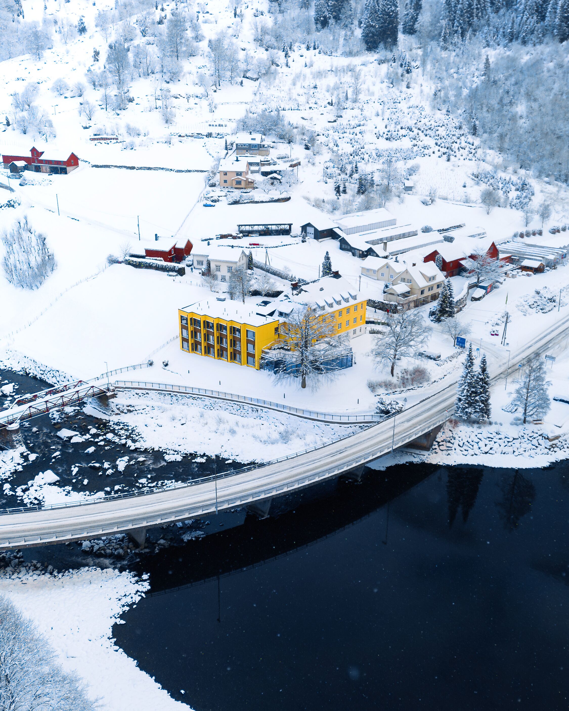 A snow-covered landscape with a bridge over a river, buildings, and a yellow structure.