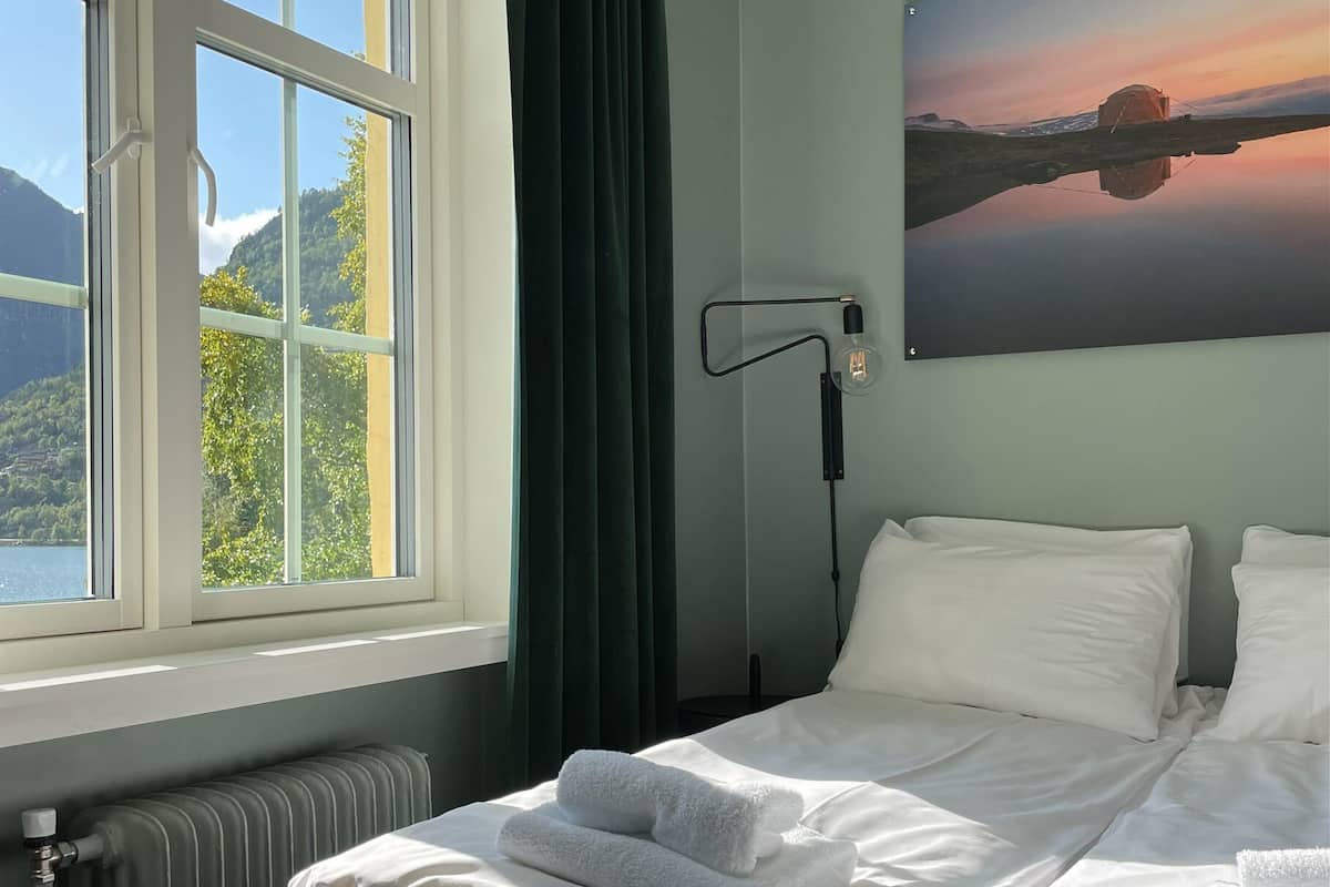 A hotel room with a large window, a bed with white linens, a radiator, and a painting of a sunset.