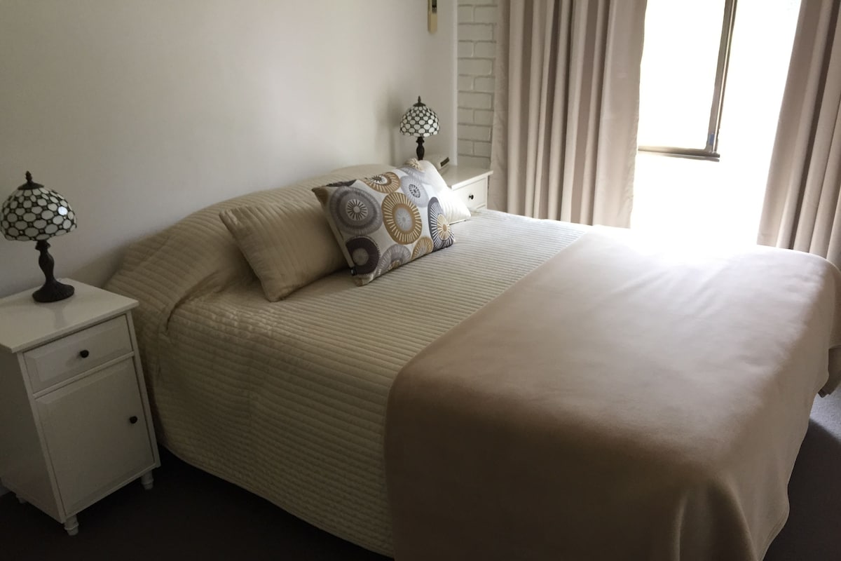 Una cama bien tendida con una colcha color beige y almohadas estampadas, una mesita de noche con una lámpara y una ventana con cortinas.