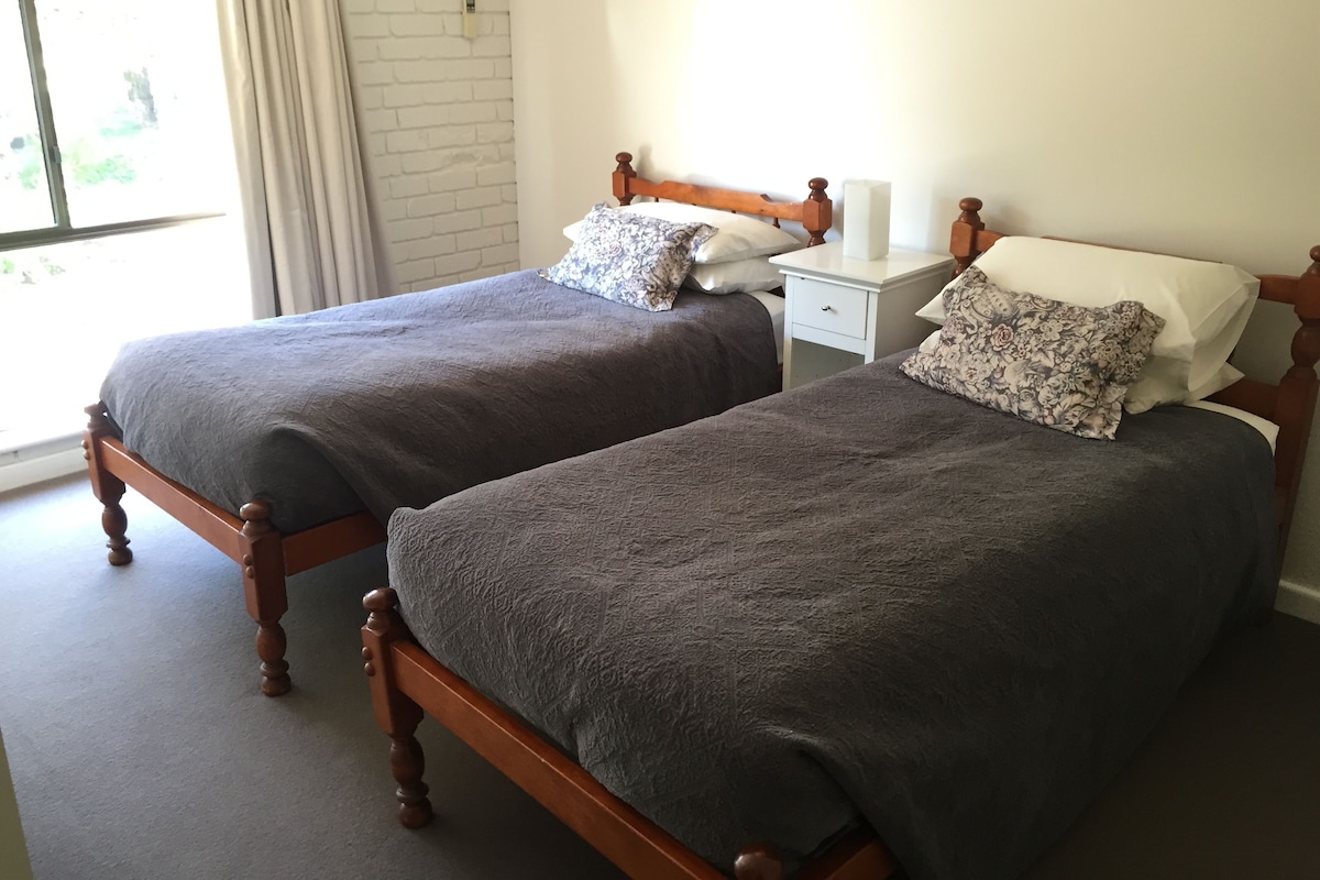 Dos camas individuales con cabeceras de madera y ropa de cama gris oscuro, una mesita de noche blanca con lámpara, y una ventana con cortinas.