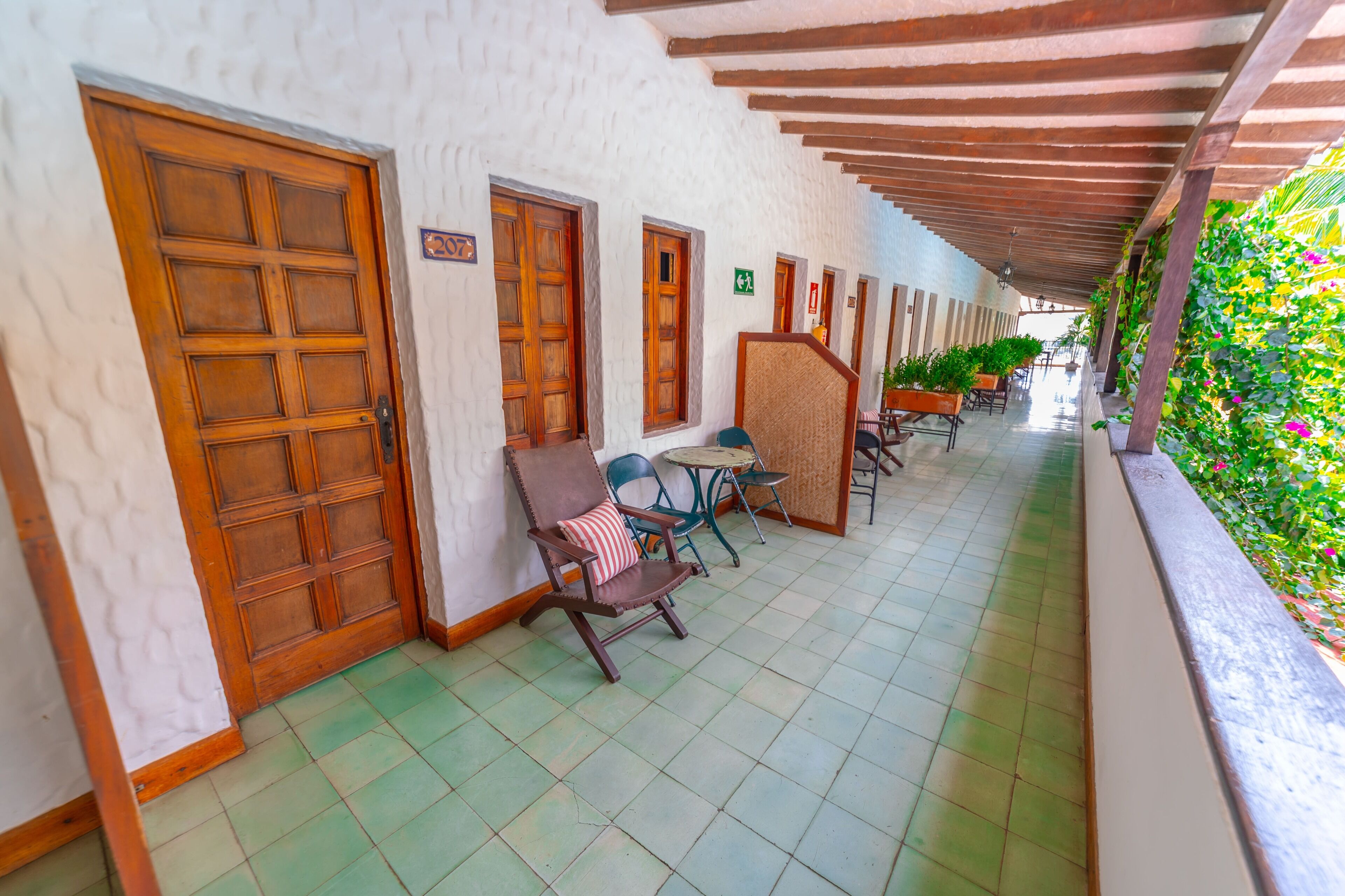 A corridor with wooden doors, a covered area with chairs and tables, and a green tiled floor.