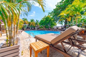 Un espace aménagé au bord de la piscine, avec des chaises longues et une table en bois, entouré de palmiers et surplombant une piscine à l’eau d’un bleu limpide.