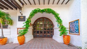 A hotel entrance with a tiled pathway, a wooden door, and a decorative arch of green vines.