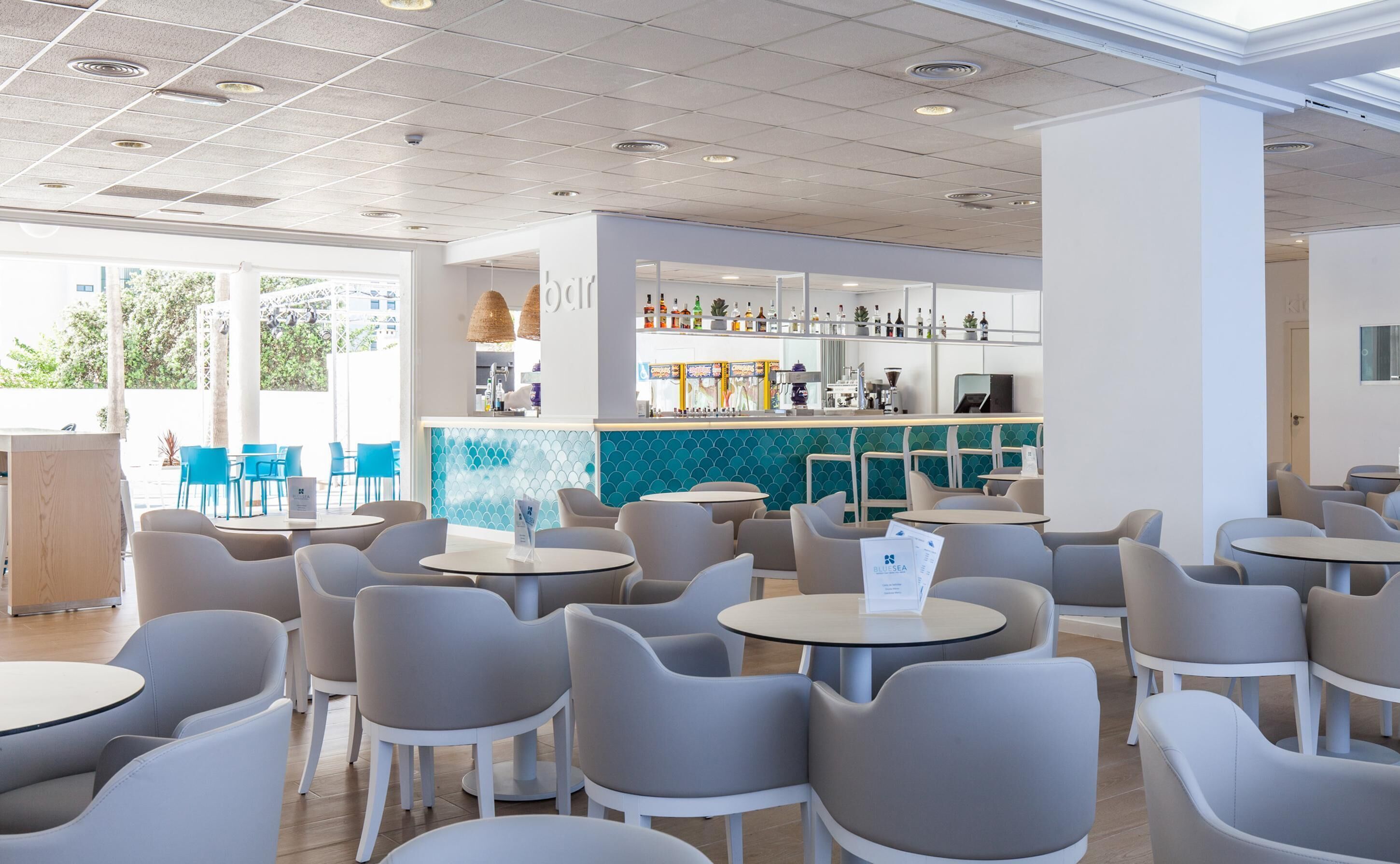 Un café moderne avec des tables rondes et des chaises grises, un bar avec des étagères remplies de bouteilles, et une vue sur la verdure à l’extérieur.
