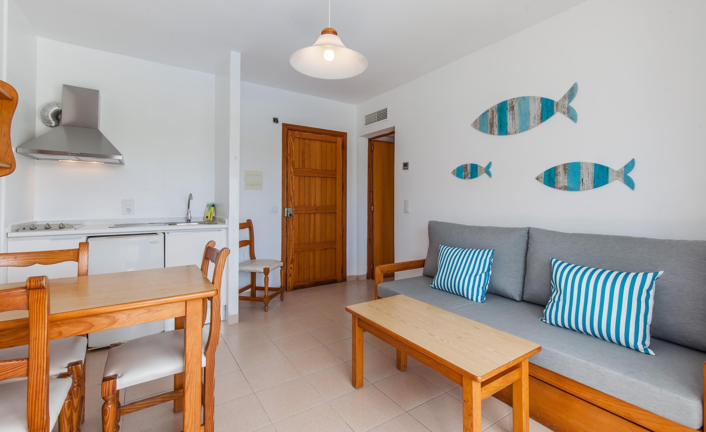 Una sala moderna con un sofá gris, almohadones a rayas, una mesa de comedor de madera y sillas. Se observa un área de cocina con electrodomésticos blancos y una puerta de madera.