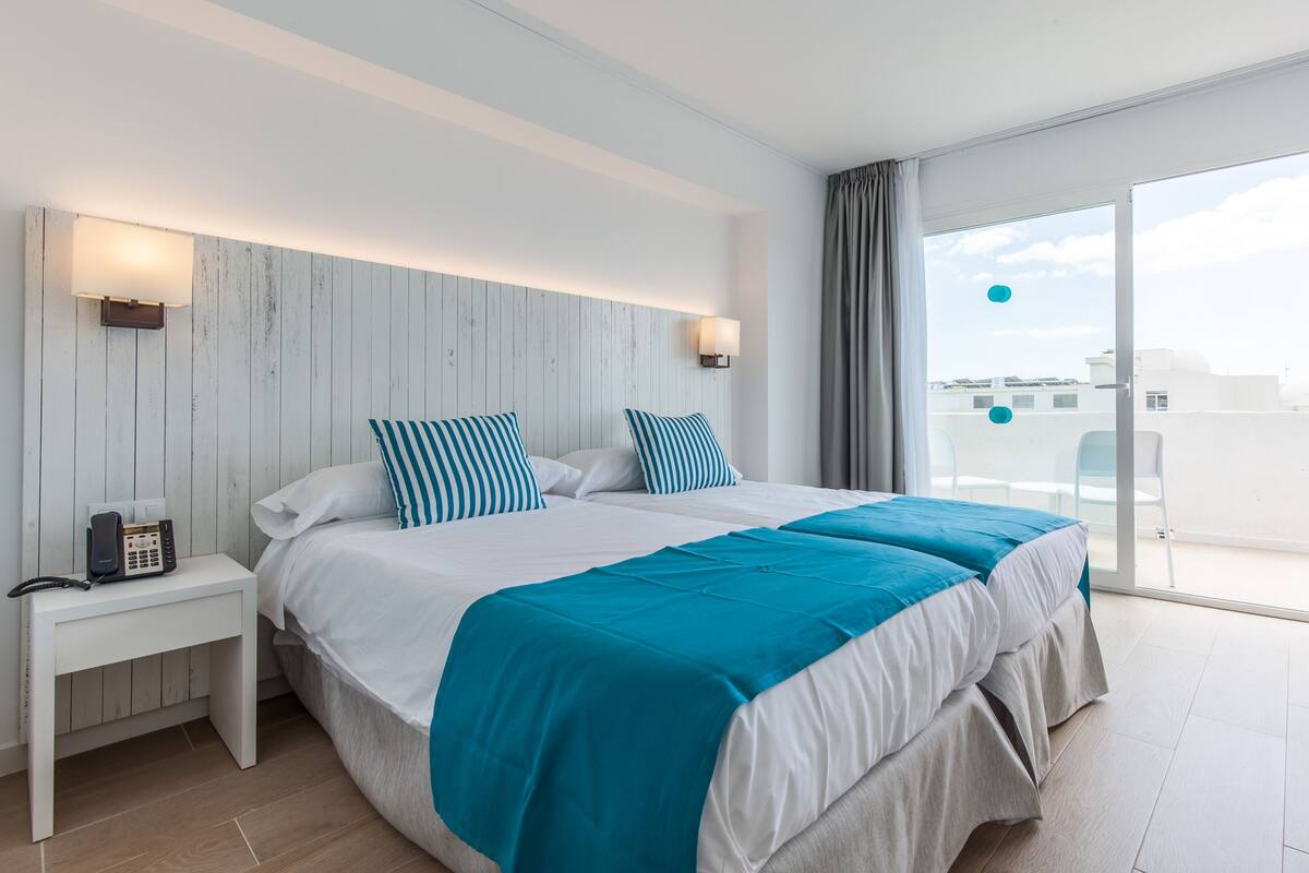 A modern hotel room with a large bed, two bedside tables, a telephone, and a view of the city through large windows.