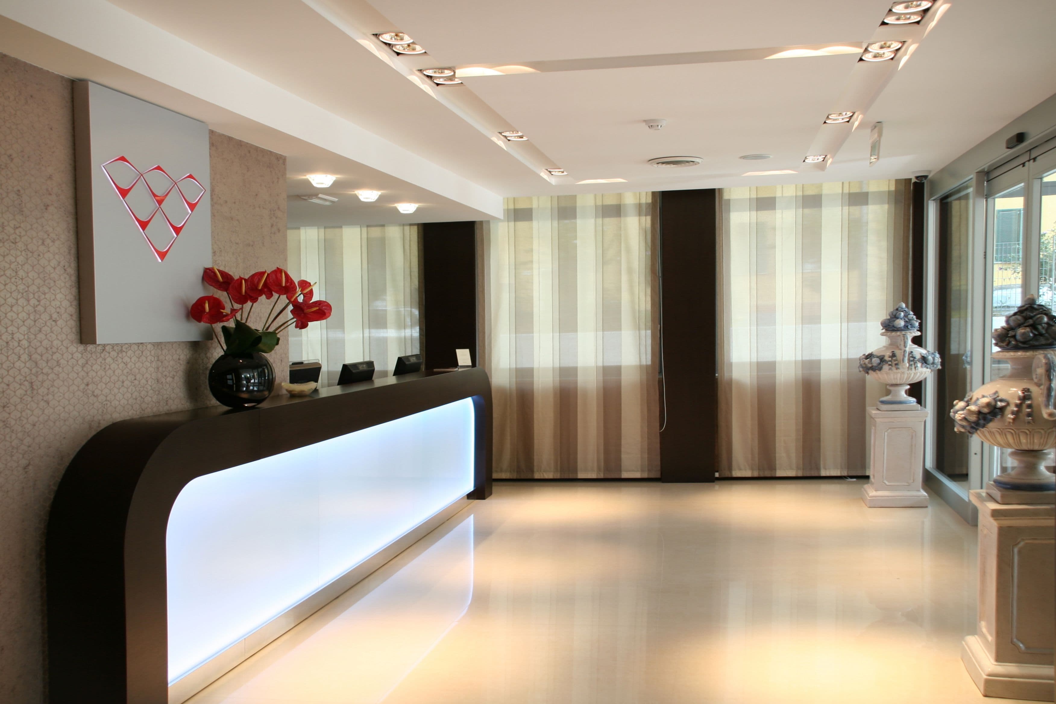 A modern reception area with a curved reception desk, a vase of red flowers, a decorative wall piece, and a large window with striped curtains.