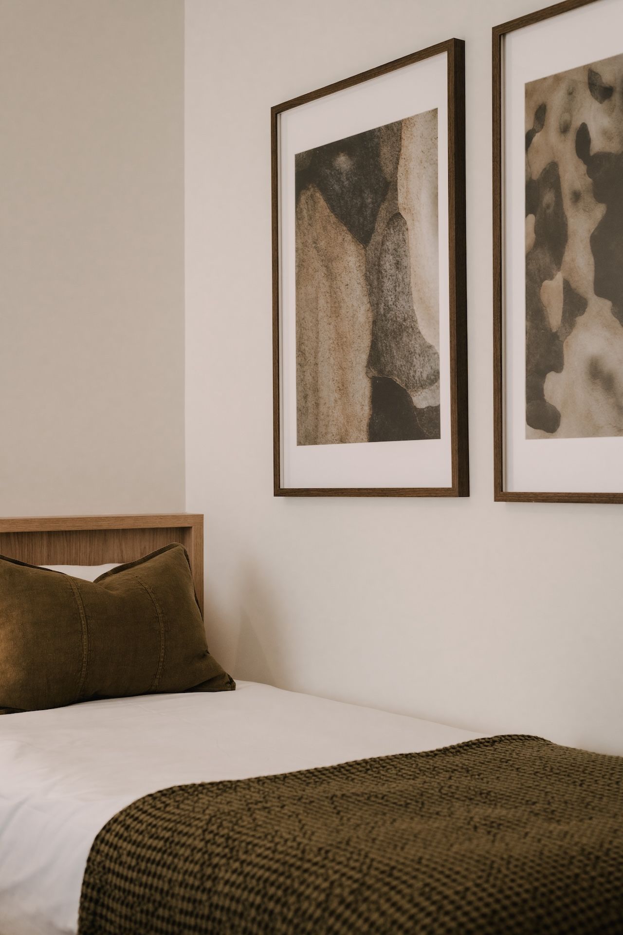 A neatly made bed with a wooden headboard, two framed abstract paintings on the wall, and a brown throw blanket on the bed.