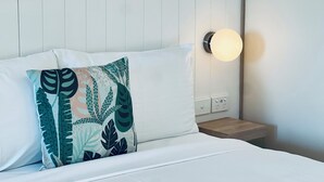 Un lit bien fait, avec du linge de lit blanc, un coussin décoratif orné d’un motif de feuilles et un luminaire fixé au mur.