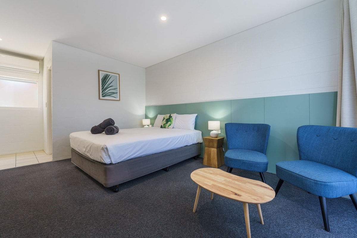 A hotel room with a bed, two blue armchairs, a small wooden table, and a nightstand with a lamp.