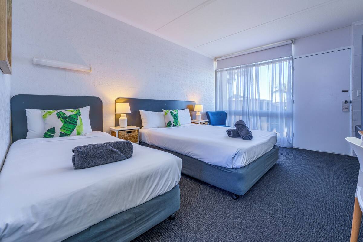 A hotel room with two beds, a window with sheer curtains, a bedside table with a lamp, and a small chair.