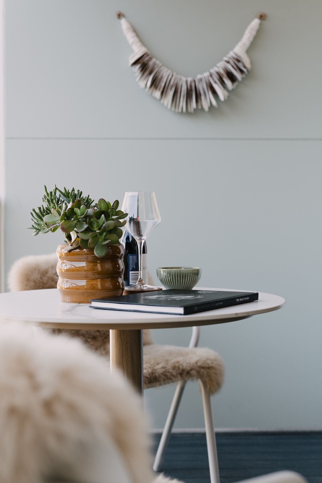 A round table with a potted plant, a wine glass, a bowl, and a book titled 'The Rock Oyster'.