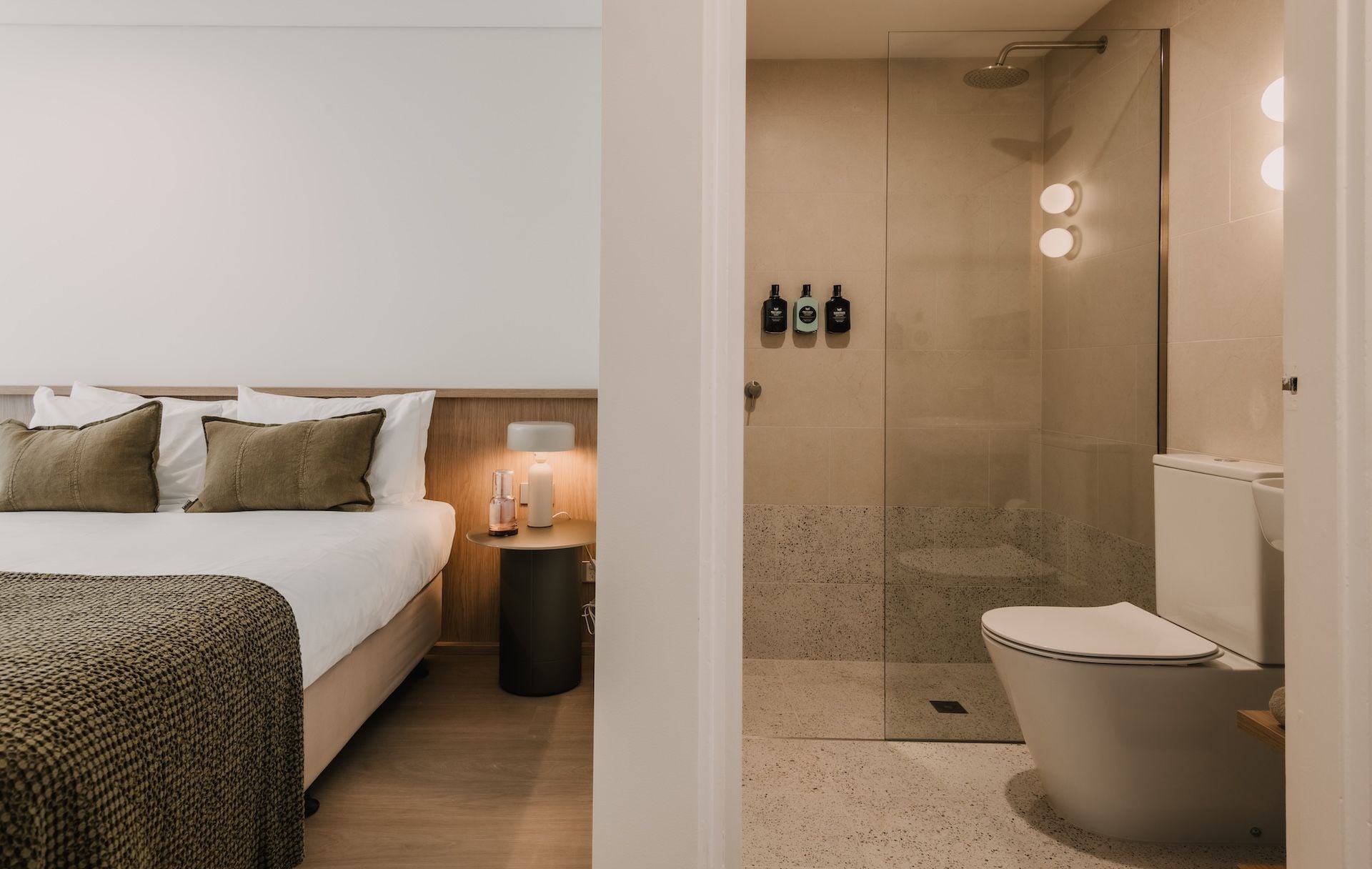 A hotel room with a bed, bedside table with a lamp, and a shower area with a toilet.
