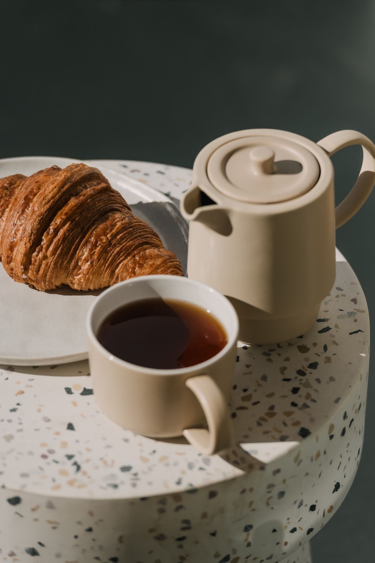 Un croissant, una tetera y una taza de té sobre un plato de cerámica con motas.