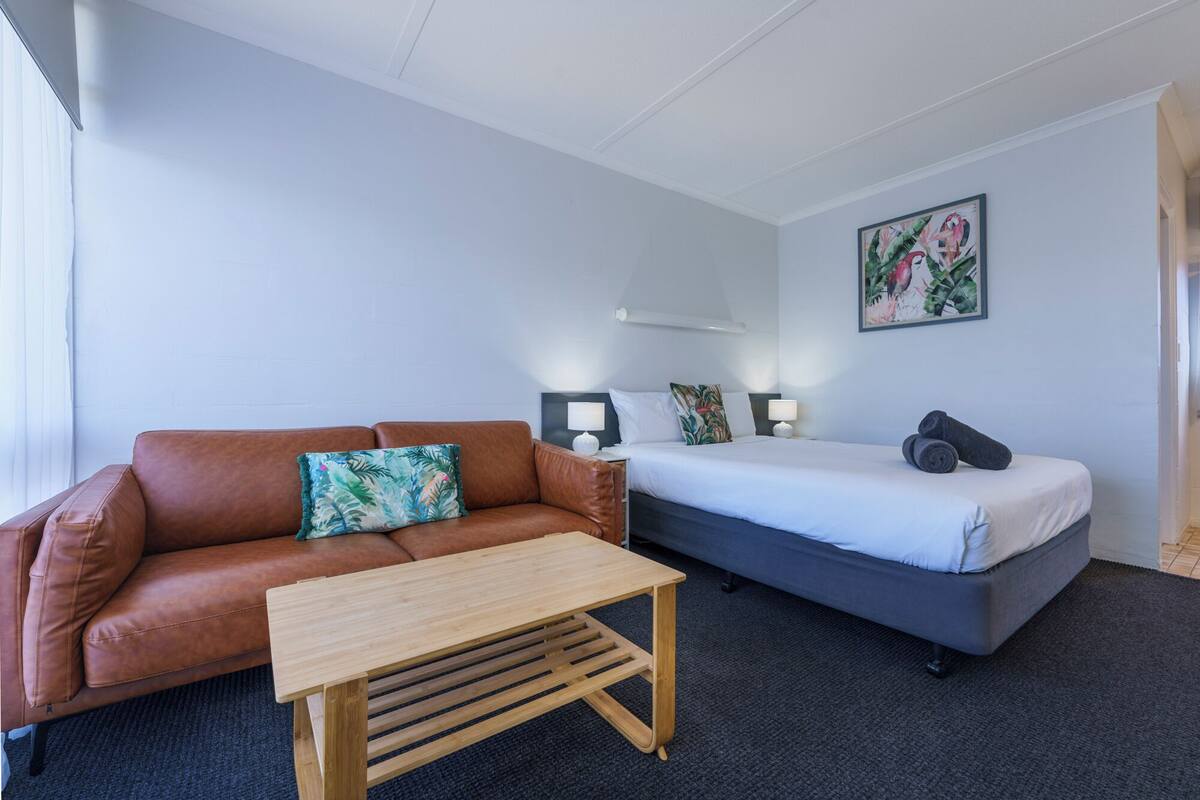 A hotel room with a brown leather sofa, a wooden coffee table, a bed with a grey headboard, and a wall-mounted painting.