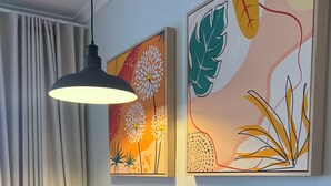 Une table ronde en bois, accompagnée d’une chaise en osier, d’une suspension et de deux tableaux abstraits représentant des motifs floraux, qui ornent le mur.