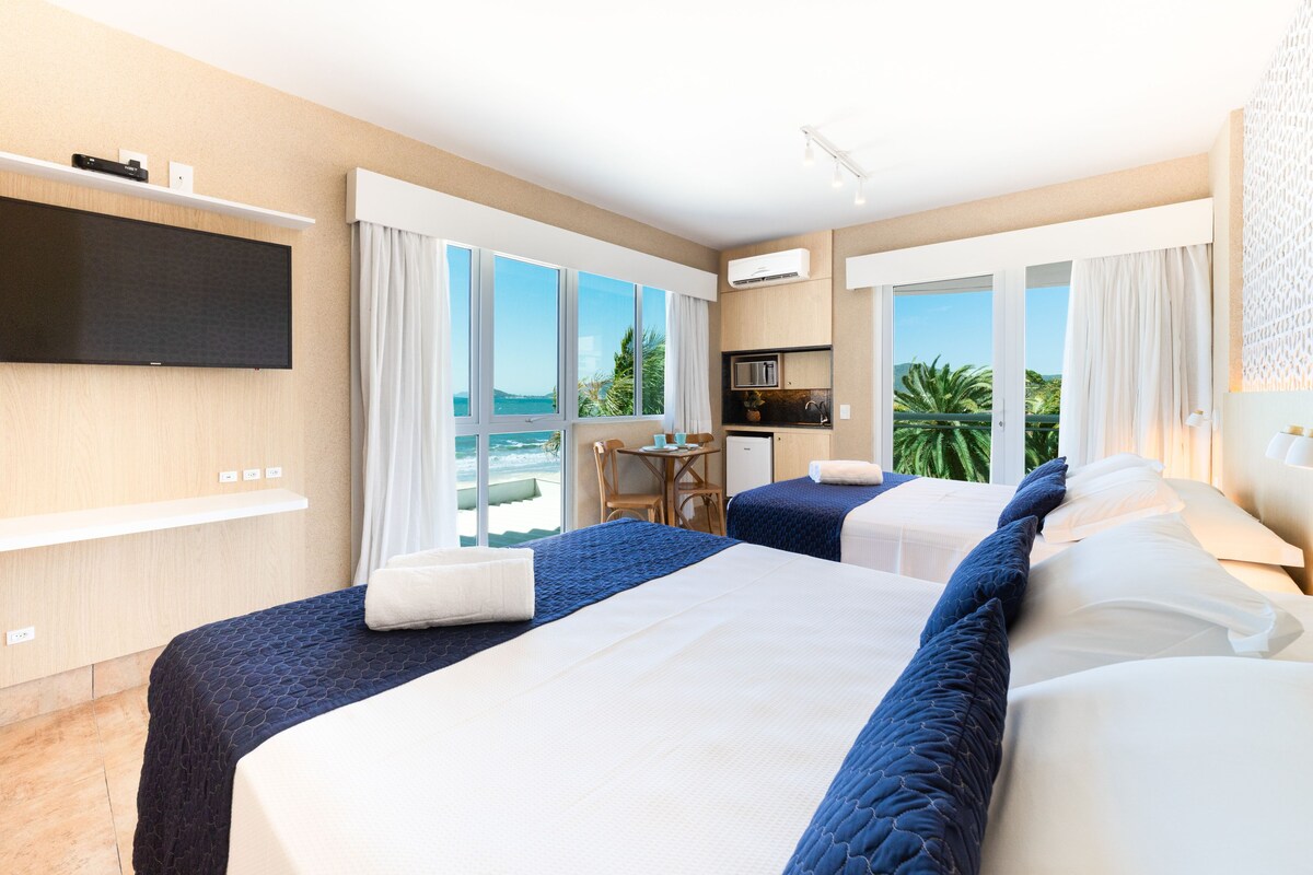 A hotel room with two beds, a TV, a desk, and a view of the sea.