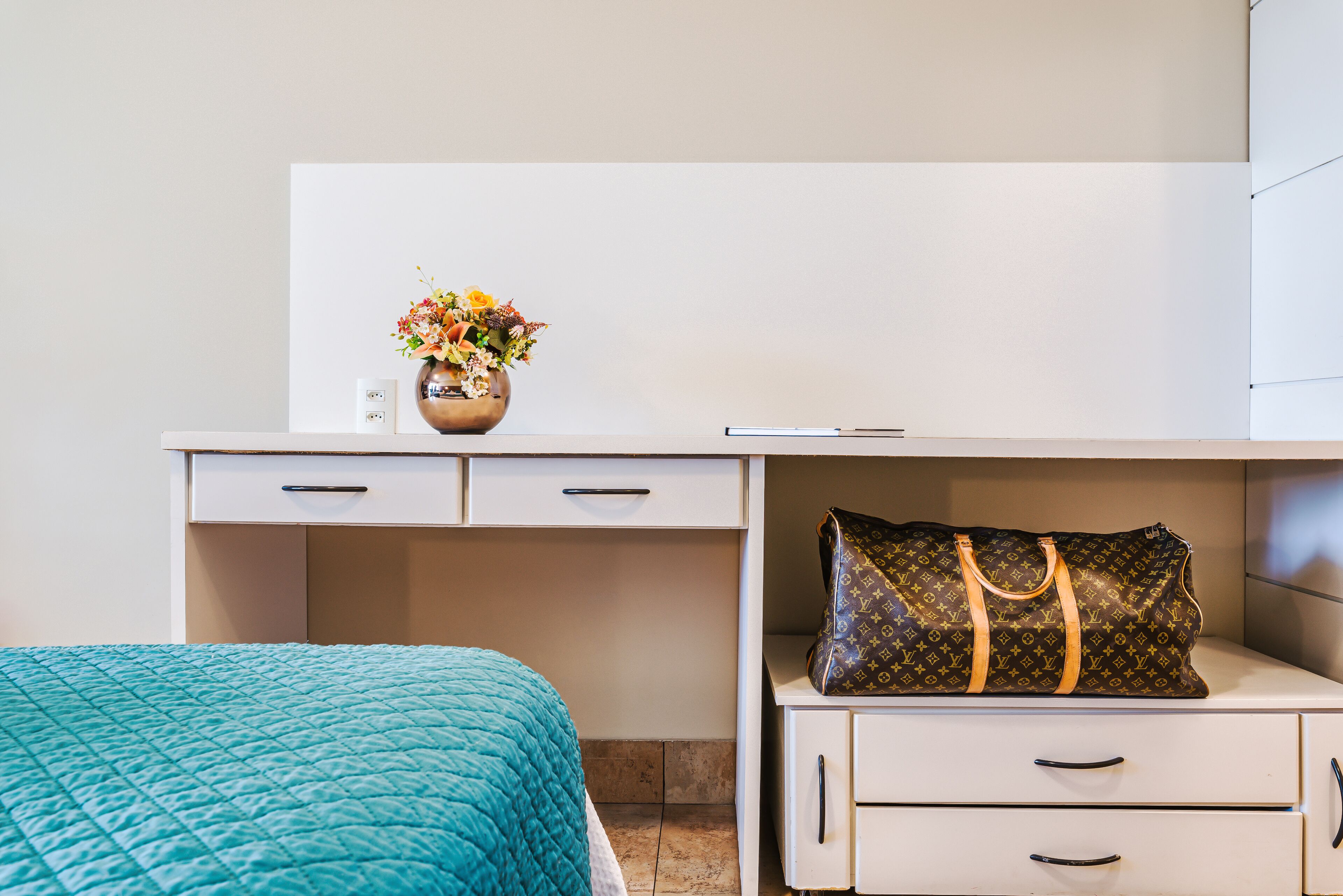 A neatly arranged bedroom with a bed, a bedside table with a Louis Vuitton bag, a vase with flowers, and a white desk with drawers.