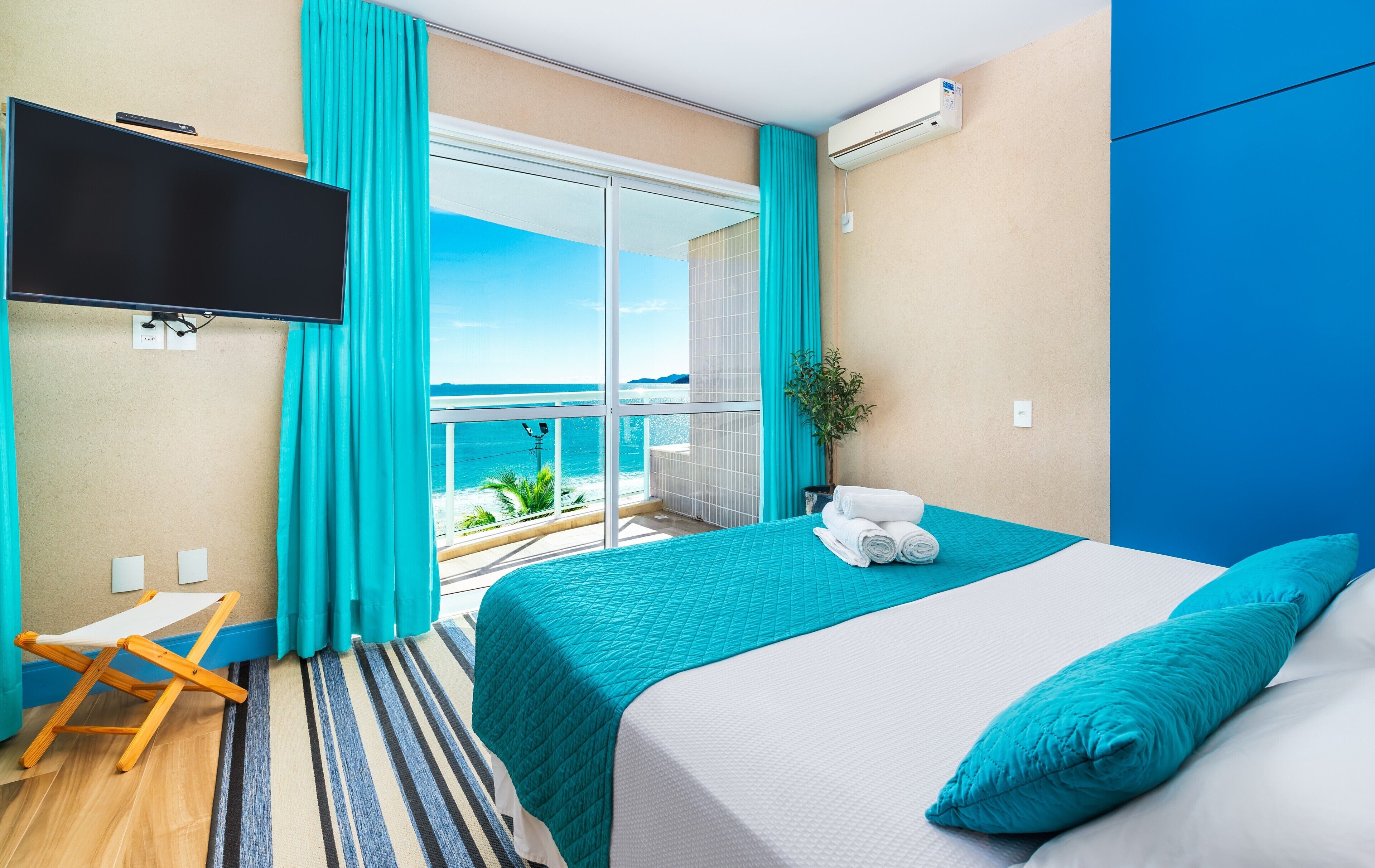 Quarto de hotel com varanda, cama com roupa de cama verde-água, televisão montada na parede e planta no parapeito da janela.
