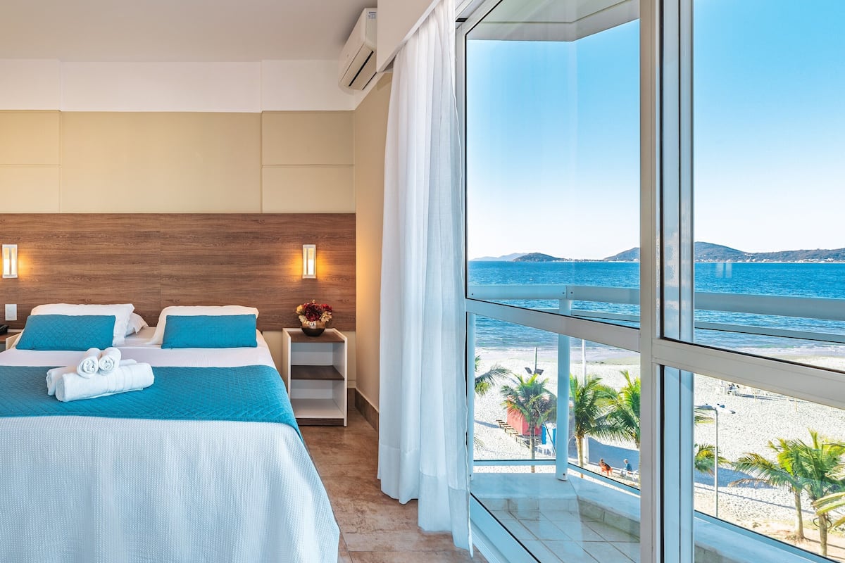 Une chambre d’hôtel avec une grande fenêtre donnant sur une plage, un lit avec une literie bleue et une table de chevet ornée d’un vase de fleurs.
