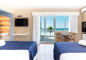 A hotel room with two beds, a TV, a small table with chairs, and a window with a view of the sea.