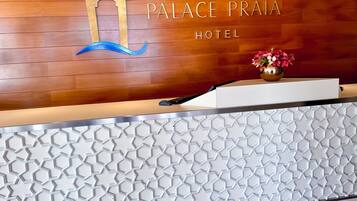 Una recepción de hotel con un fondo de paneles de madera, un gran logotipo con la letra 'P' y un jarrón con flores.