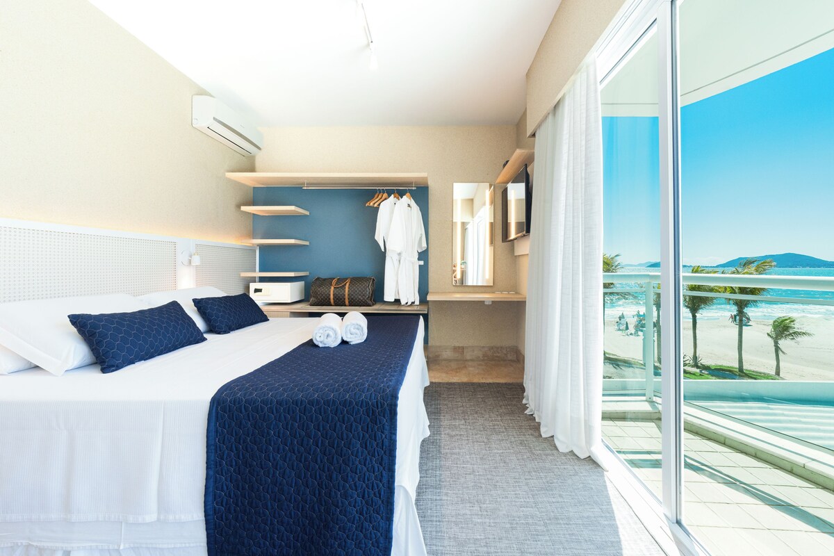 A hotel room with a bed, a seating area with a view, and a large window overlooking a beach.