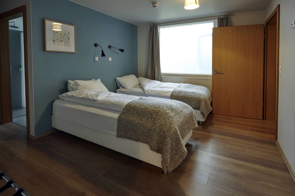 Un dormitorio con dos camas, una ventana con cortinas, una puerta y una lámpara fijada a la pared.