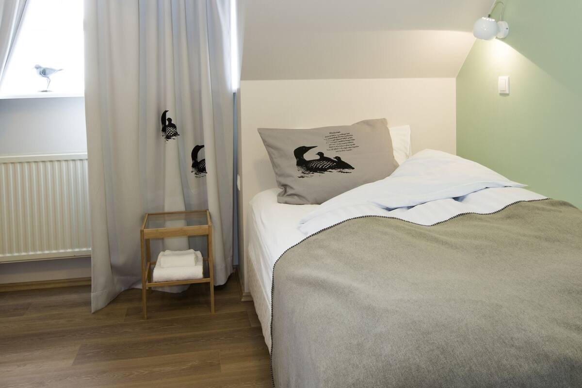 Un dormitorio con cama, una silla, una ventana con cortinas y una lámpara fijada a la pared.
