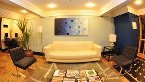 A lobby with a cream-colored sofa, two armchairs, a coffee table with magazines, a large abstract painting, and a potted plant.