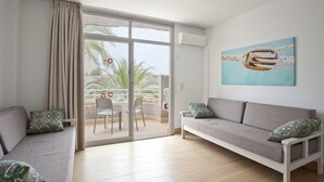 A modern living room with a sofa, a dining table, and chairs. There is a sliding glass door leading to an outdoor area with a view of palm trees. - BJ Apartamentos Club Sa Coma (Sant Llorenc des Cardassar)