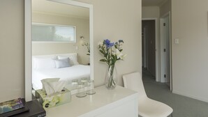 A hotel room with a bed, a desk with a vase of flowers, a mirror, and a bedside table with tissues and glasses. - Seascape Villa Bed & Breakfast (Tauranga)
