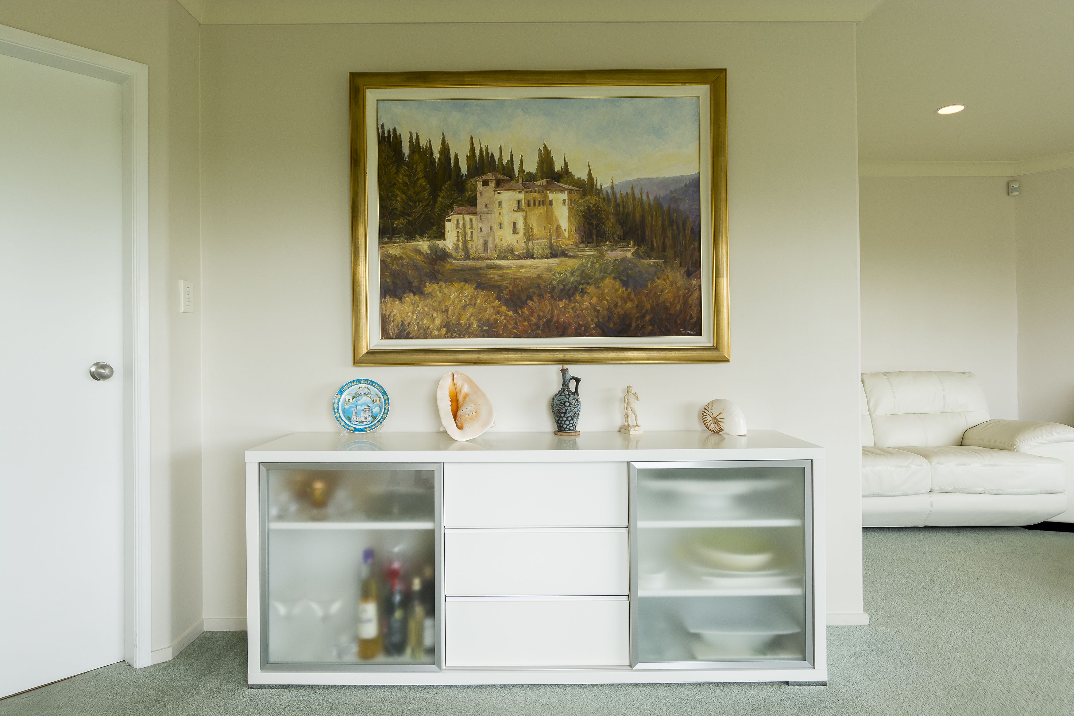 Un mueble blanco con puertas de vidrio y un cuadro de una casa en la pared.
