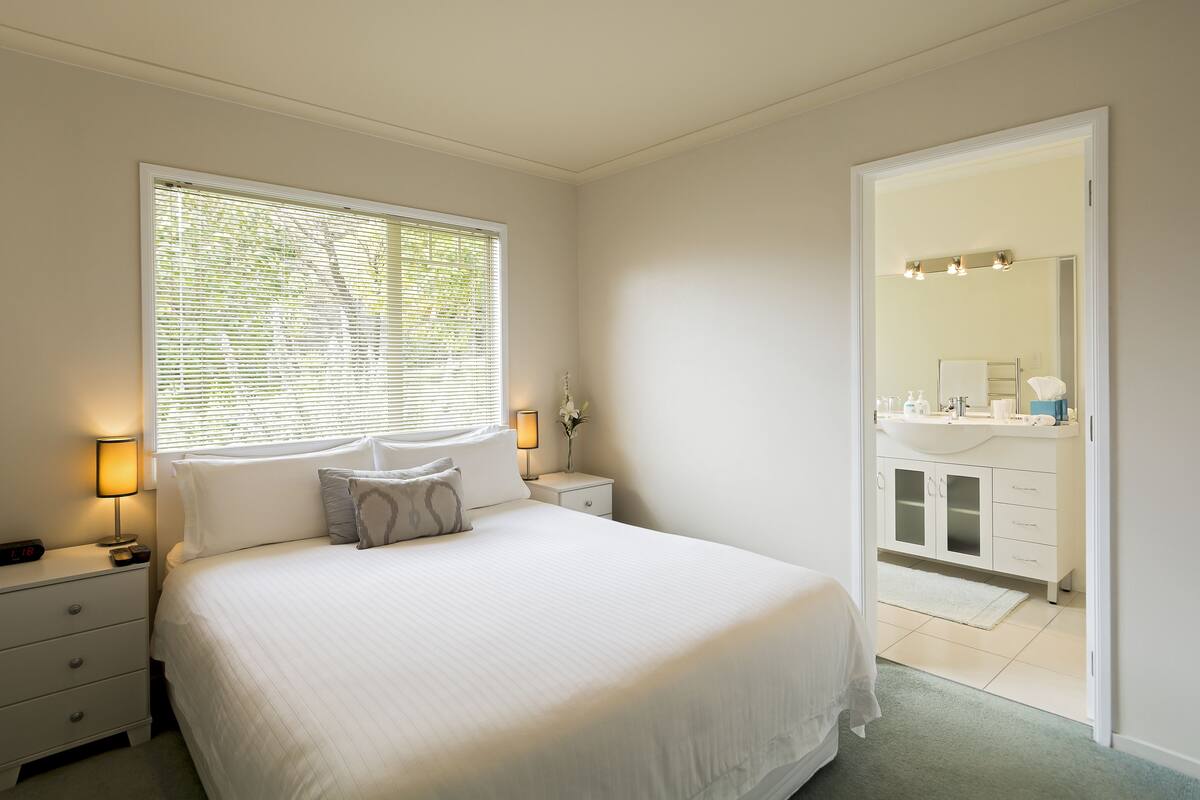 Een slaapkamer met een groot bed, nachtlampjes, een nachtkastje, een raam met zonwering en uitzicht op de badkamer.