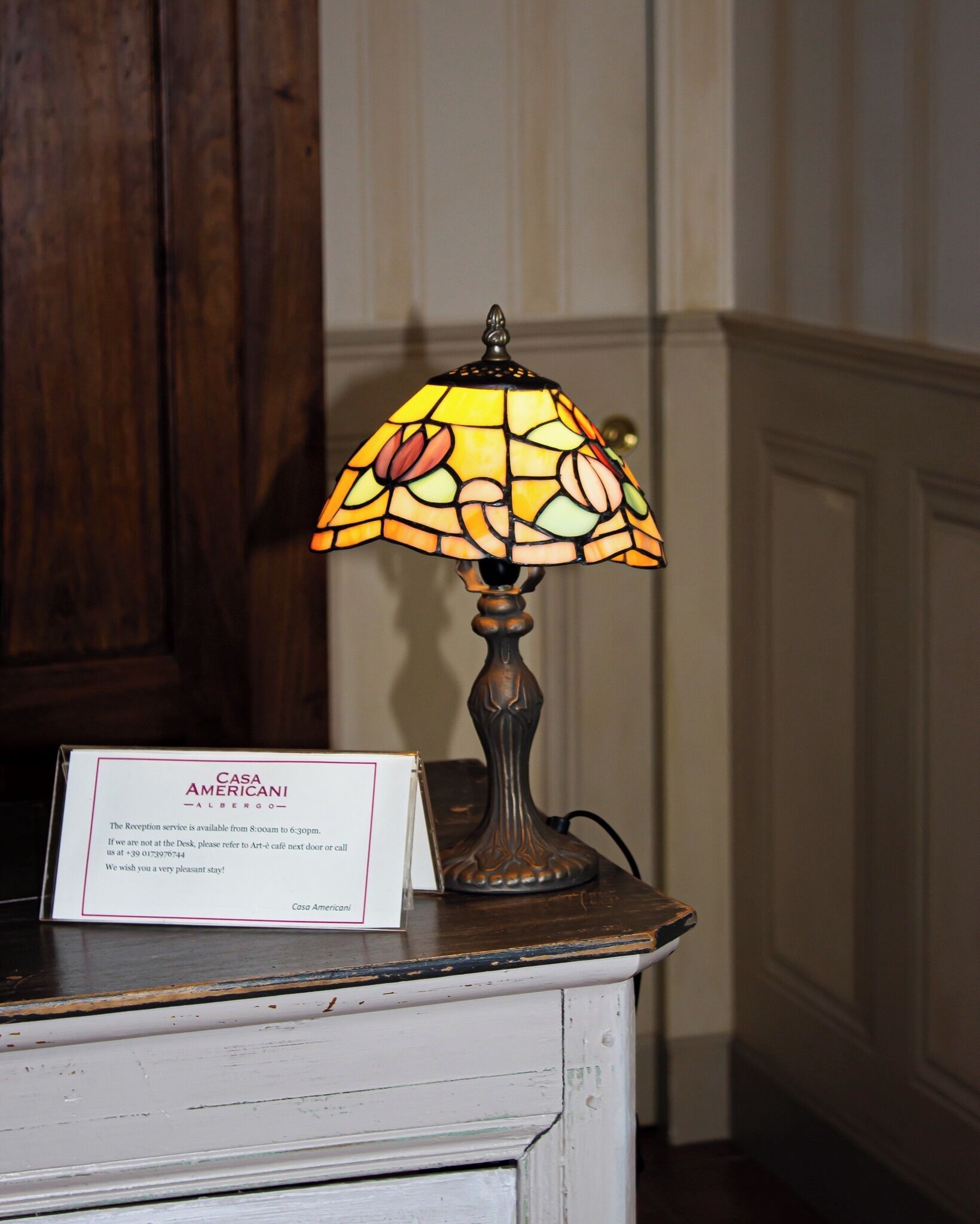 A stained glass lamp with floral patterns on a wooden table, a sign with hotel information, and a wooden cabinet in the background.
