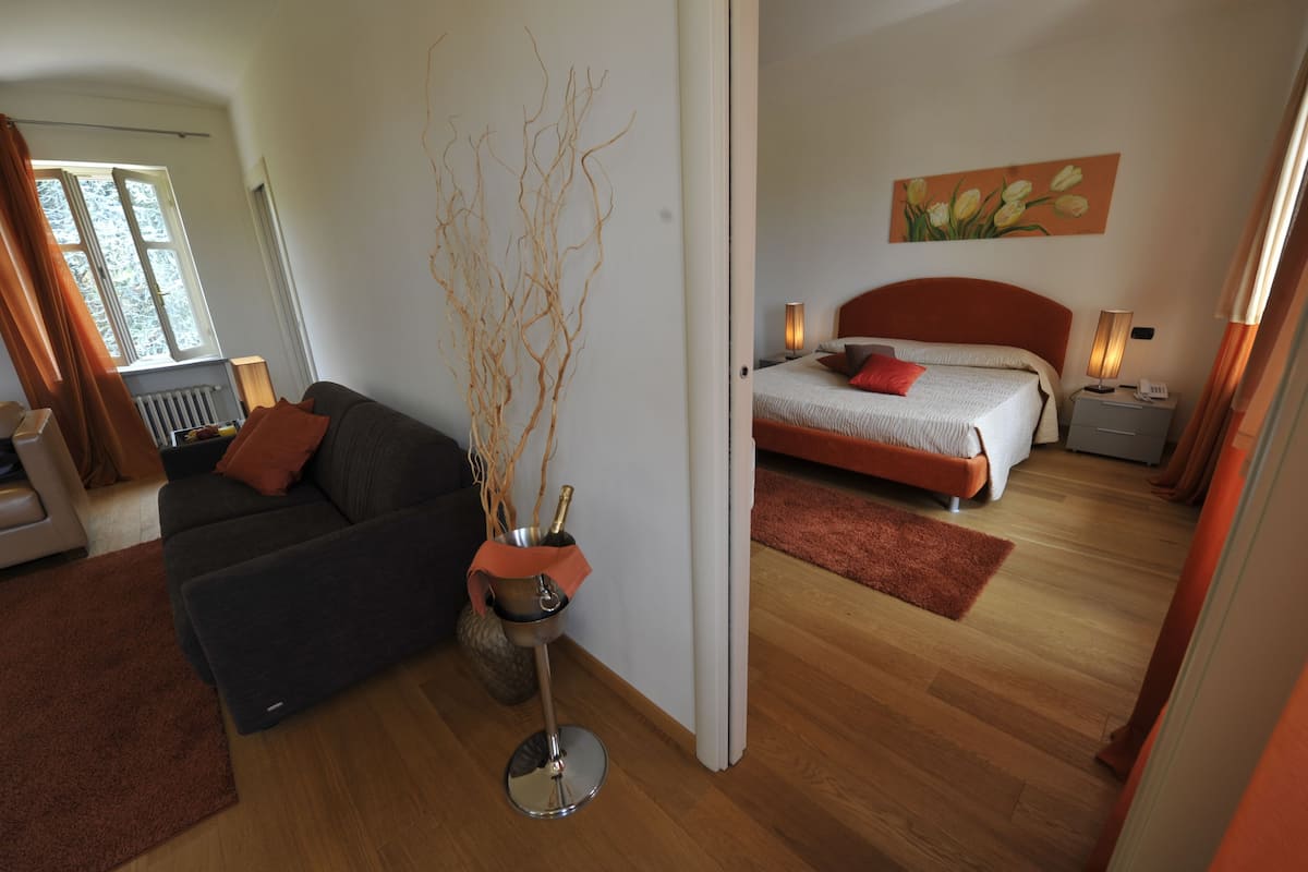 A modern hotel room with a sofa, a bedside table with a lamp, a bed with a red headboard, and a painting on the wall.