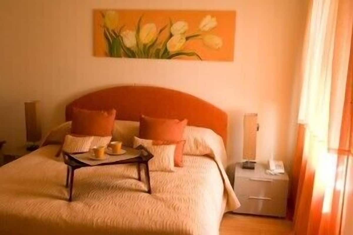 A hotel room with a bed, two pillows, a bedside table, a lamp, and a painting of flowers on the wall.