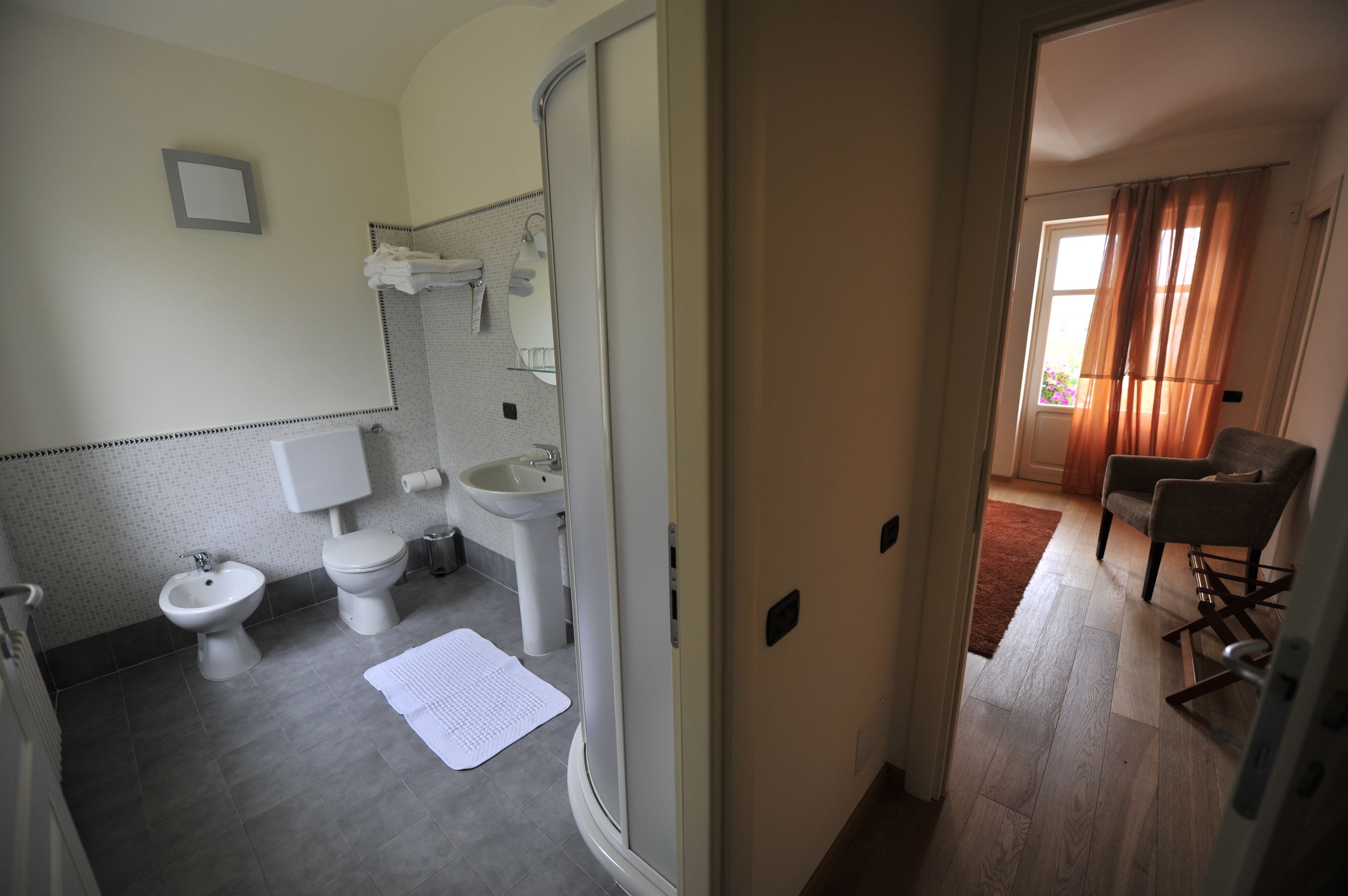 Un bagno con toilette, bidet, lavabo e specchio. È presente un portascopino con asciugamani e un tappetino bianco sul pavimento.