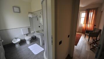 Ein Badezimmer mit Toilette, Bidet, Waschbecken und Spiegel. Handtücher und eine weiße Matte liegen auf einem Handtuchhalter und dem Boden.