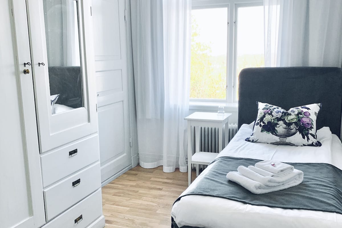 A bedroom with a bed, a white wardrobe, a window with curtains, and a small table.