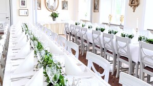 A long banquet table with white tablecloths, glassware, and floral centerpieces, set in a well-lit room with white chairs and a chandelier. - Drottning Victorias Hotell och Vilohem (Borgholm)