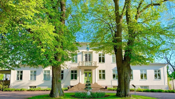 A two-story white building with a central entrance, surrounded by a gravel driveway and lush greenery. - Drottning Victorias Hotell och Vilohem (Borgholm)