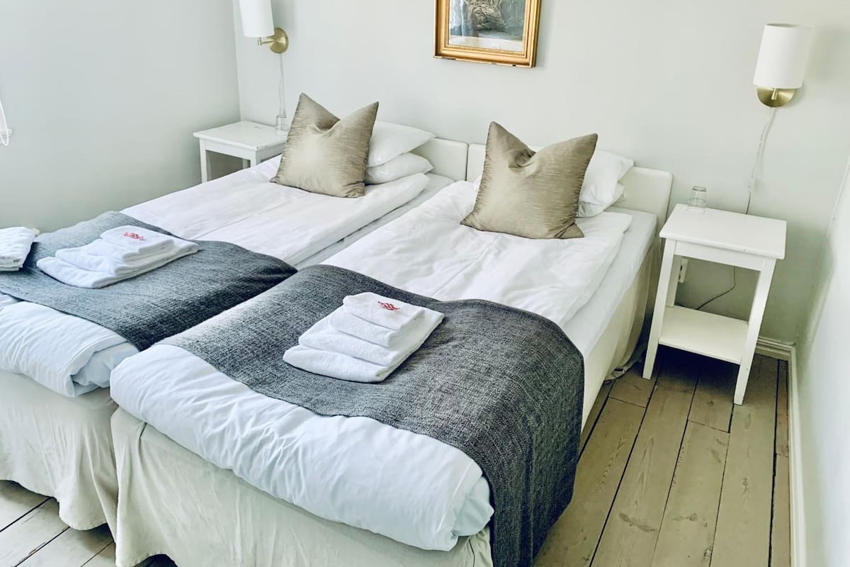 A hotel room with two beds, white bedding, grey and beige pillows, a framed artwork on the wall, and bedside lamps.